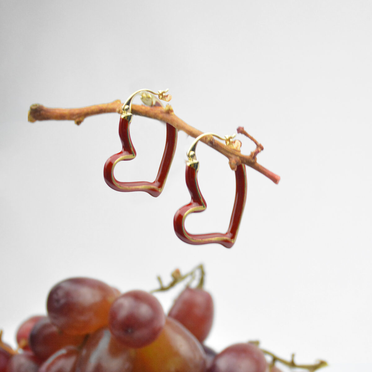 Heart-shaped earrings featuring a red design with gold accents, hanging from a branch, surrounded by grapes.
